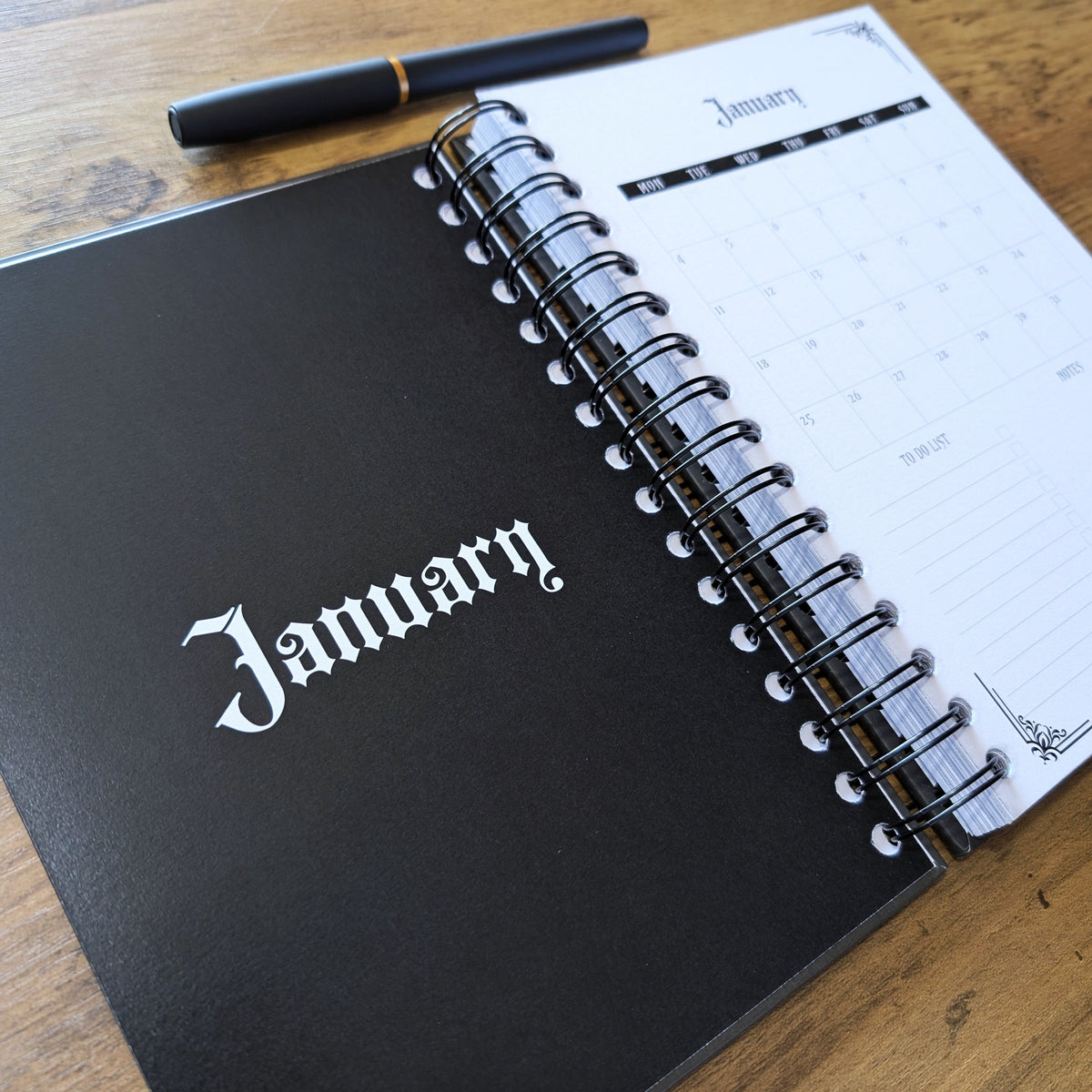 A black spiral-bound January planner or notebook lies open on a wooden table, with a black pen resting above it. The cover shows the word January in white gothic lettering, and the open page shows a monthly calendar layout.
