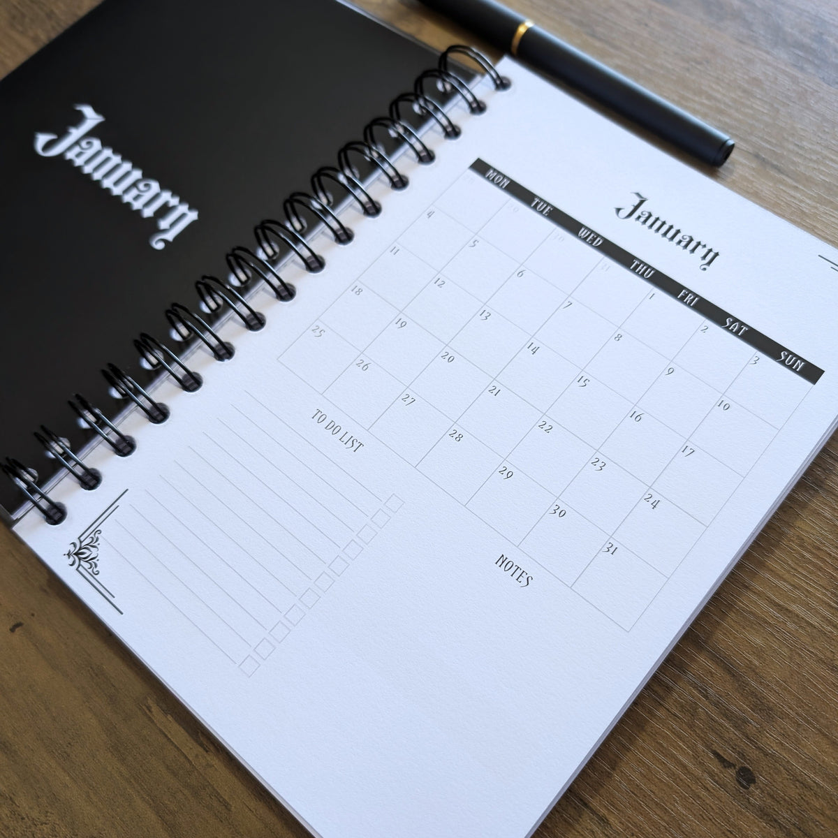A spiral-bound January desk calendar with a black cover and white calendar pages, shown open on a wooden tabletop with a black pen resting on top.