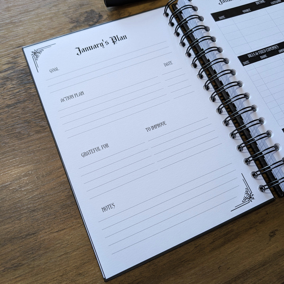 An open spiral-bound planner on a wooden table, showing the January's Plan page with lined writing areas and a black pen resting above it.