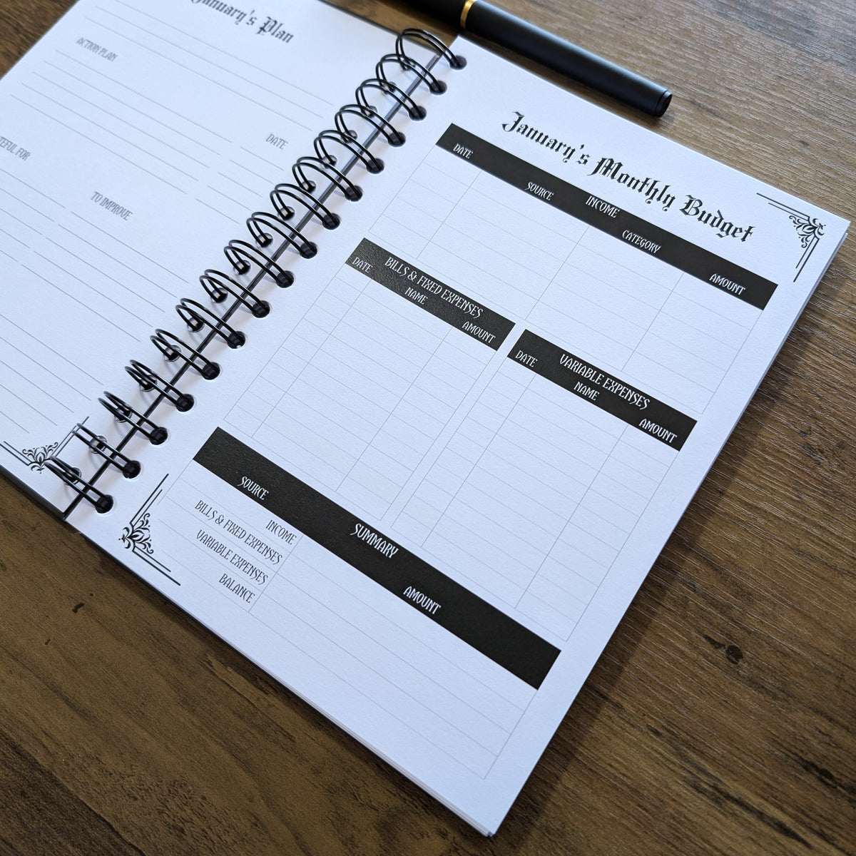 A spiral-bound monthly budget planner notebook open on a wooden table, with a black pen resting across the top page.