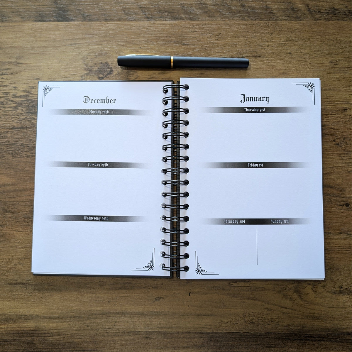 An open spiral-bound planner or agenda on a wooden tabletop, showing blank pages for December and January with a black pen placed above it.