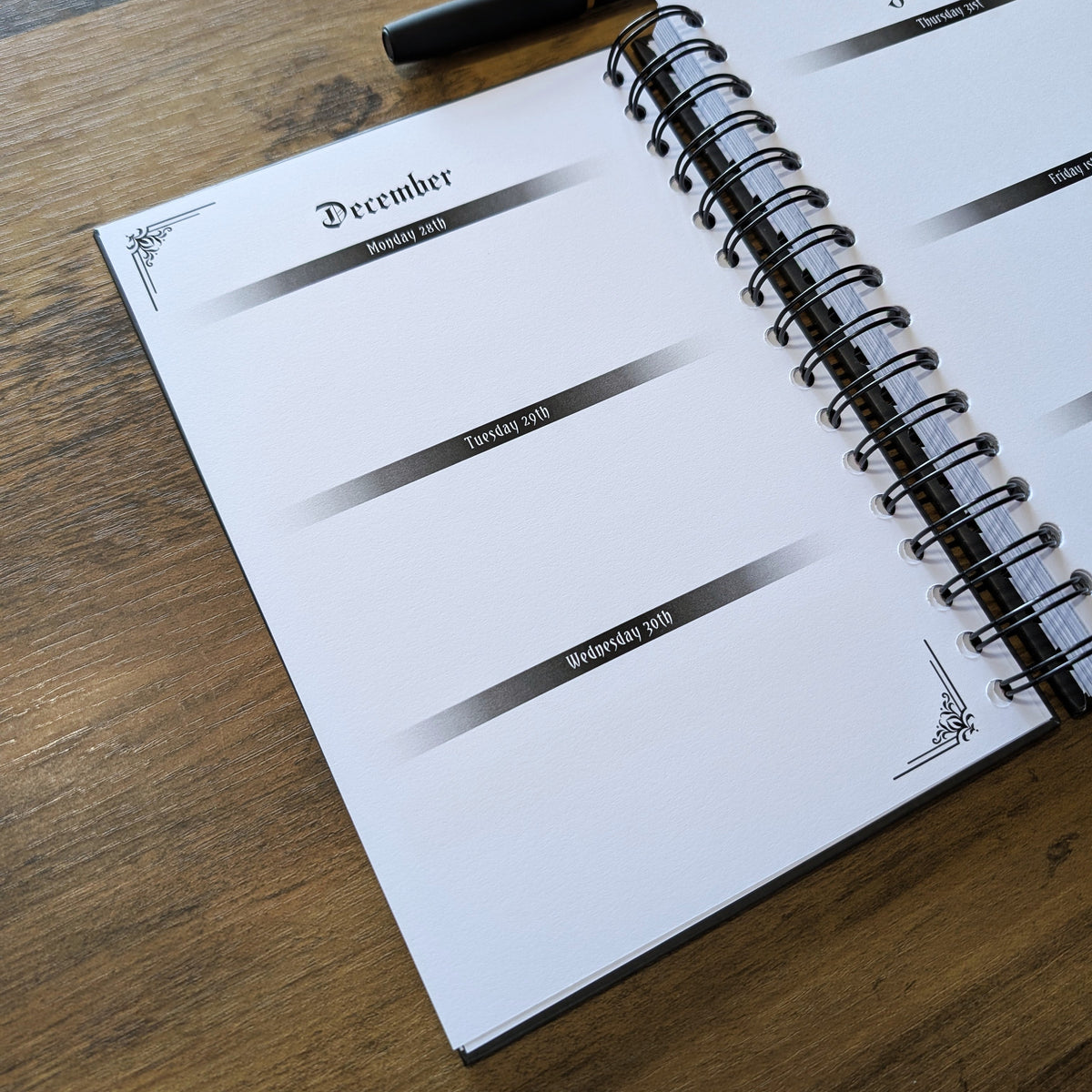 An open spiral-bound planner or notebook on a wooden table, showing blank weekly pages for December, January, and February. A black pen with a gold band rests across the top.