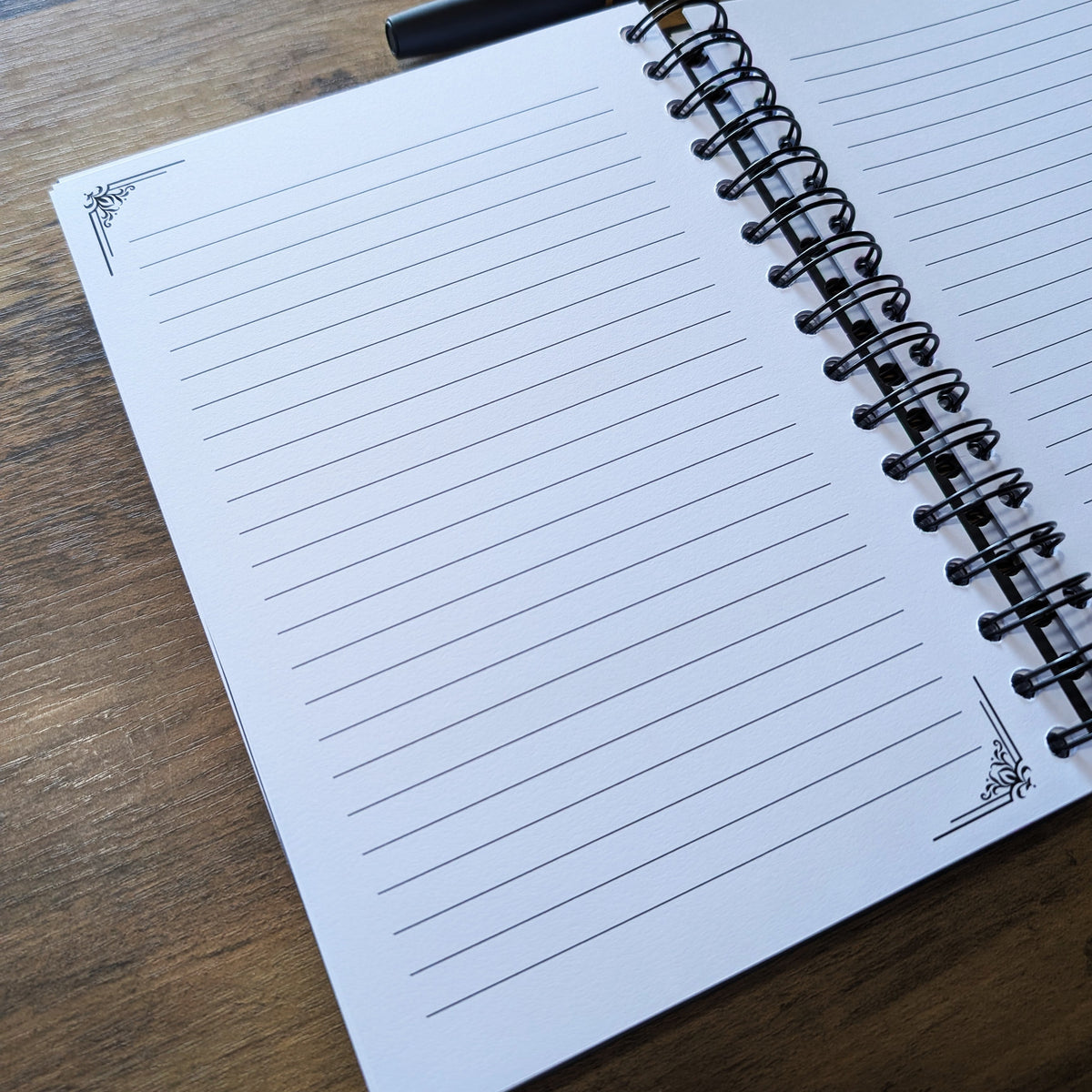 An open spiral-bound lined notebook on a wooden tabletop, with a black pen resting across the top page.