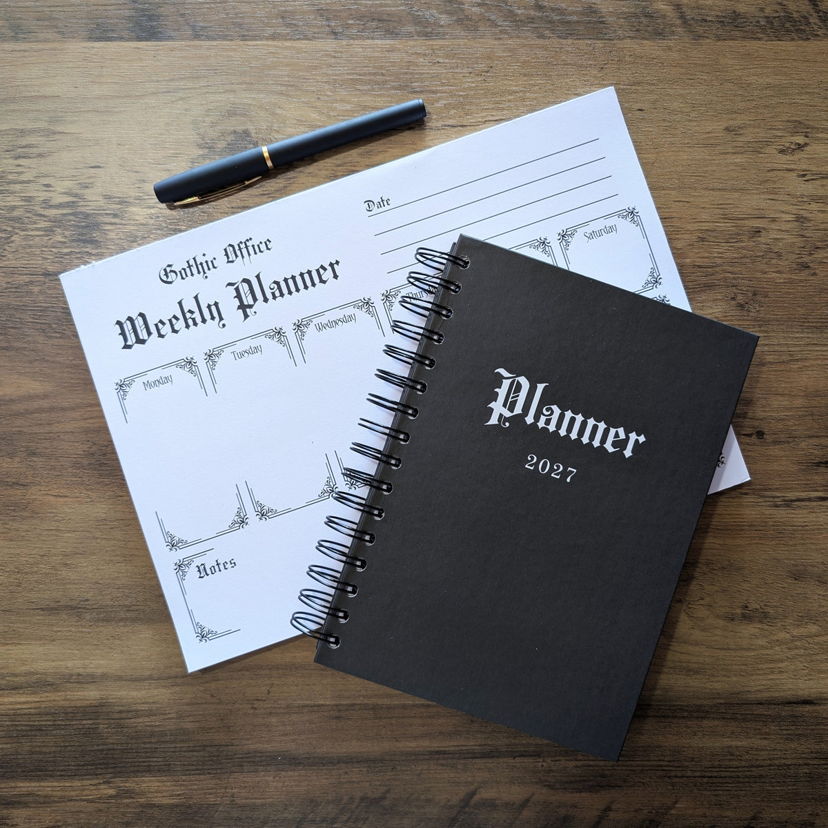 A black spiral-bound planner labeled 'Planner 2027' lies on a wooden tabletop, partially covering a white weekly planner notepad and a black pen with a gold band.